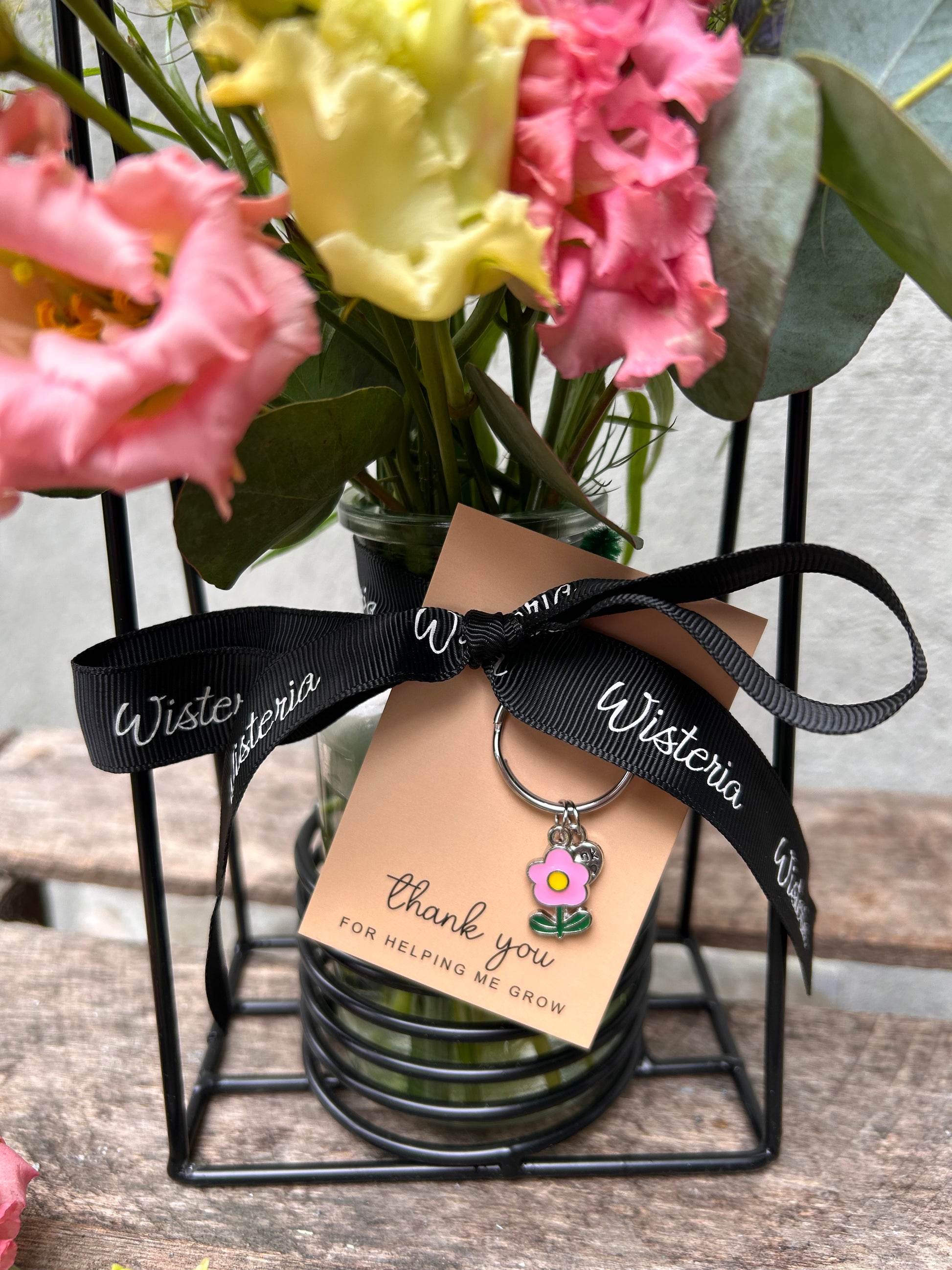 A charming clear glass bottle filled with fresh seasonal wildflowers adorned with a delicate “Thank You for Helping Me Grow” keyring charm, styled with natural textures and whimsical elegance.